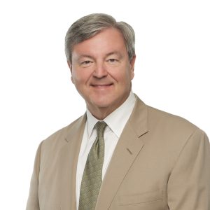 A smiling individual wearing a beige blazer and a tie against a plain background.