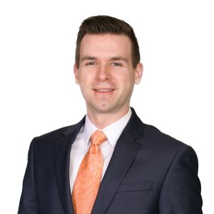 A man wearing a navy suit with an orange tie is smiling against a white background.