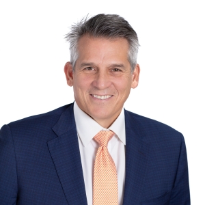 A smiling person wearing a blue suit, white shirt, and striped orange tie with a professional headshot style background.