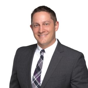 A smiling individual wearing a suit and a plaid tie against a white background.