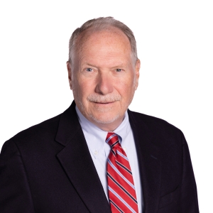 A person with a short haircut and a beard is wearing a dark suit with a red striped tie.