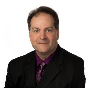 A man wearing a black suit jacket over a purple shirt with a matching tie is posing for a professional portrait against a white background.