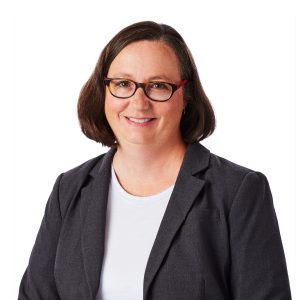 A woman with glasses wearing a blazer and white top smiles against a plain background.
