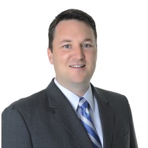 A person is smiling and dressed in professional attire with a striped tie and a jacket against a plain, light background.