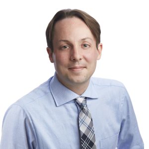 A person is dressed in business attire with a blue shirt and a gray striped tie against a plain background.