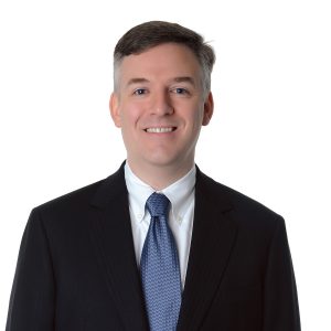 A person is wearing a dark suit, white shirt, and patterned tie, and is smiling at the camera against a white background.