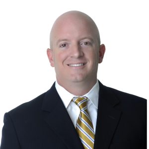 A smiling individual with a shaved head dressed in a professional black suit, white shirt, and patterned yellow tie against a white background.