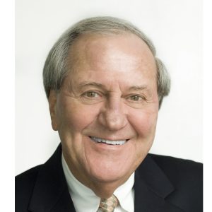 A smiling individual with a business suit and tie is shown against a white background.