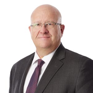 A person with a neutral expression wearing glasses, a dark suit, a white shirt, and a purple striped tie.