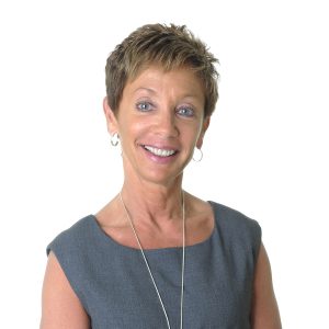 A smiling person with short hair, wearing a sleeveless top and a necklace, stands against a white background.