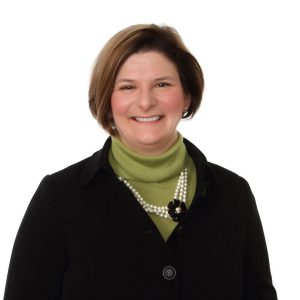 A smiling woman wearing a turtleneck, blazer, and pearl necklace against a white background.