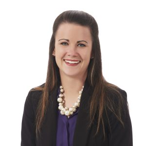 A smiling woman wearing a dark blazer and a pearl necklace against a white background.