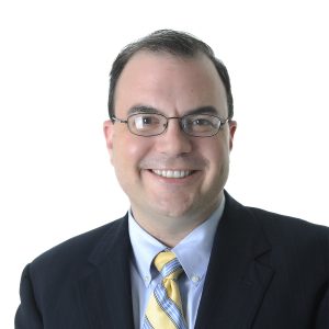 A smiling individual wearing glasses, a dark suit, white shirt, and a yellow-striped tie against a white background.