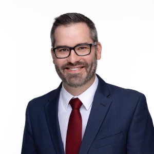 A person with glasses is wearing a blue suit, white shirt, and red tie, and is smiling at the camera.