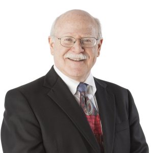 A smiling man wearing glasses, a black suit, a white shirt, and a red tie against a white background.