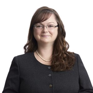 A woman with glasses is wearing a dark-colored blazer over a lighter top, with a necklace and her hair styled to one side, set against a plain background.