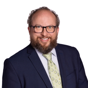 A smiling individual with a beard, wearing glasses, a blue suit jacket, and a tie with a polka dot pattern.