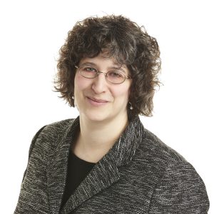 A smiling person with curly hair wearing glasses and a tweed blazer against a white background.