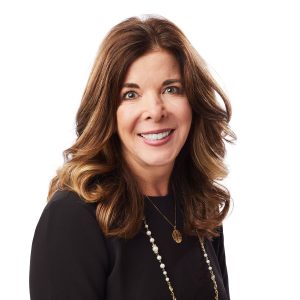 A smiling woman with long brown hair wearing a black top and a necklace with a circular pendant and long beads.