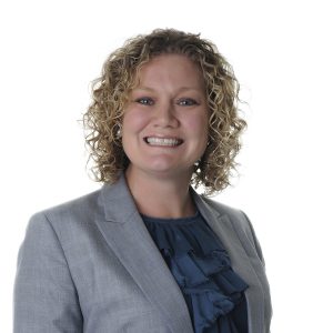 A person with curly hair is smiling and wearing a grey blazer over a blue ruffled blouse against a white background.