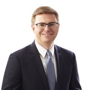 A smiling individual wearing glasses, a dark suit, and a light blue tie against a white background.