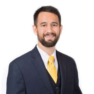 A person with a beard is wearing a dark suit and a bright yellow tie, smiling against a white background.