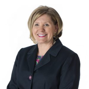 A smiling individual with short hair is wearing a dark-colored blazer over a floral blouse against a white background.