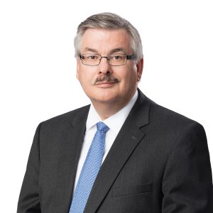 A man with a mustache is wearing a dark suit, white shirt and light blue tie against a white background.