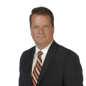 A person wearing a suit with a striped tie poses for a professional portrait against a light background.