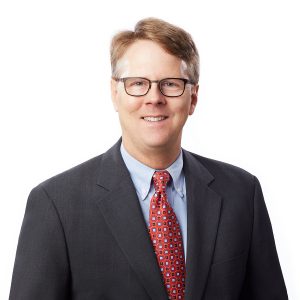 A smiling man with glasses, wearing a gray suit and a red tie with a white pattern, stands against a white background.