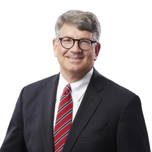 A smiling individual with glasses wearing a black suit, a white shirt, and a red striped tie against a white background.