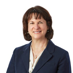 A smiling person with short hair is wearing a dark blazer over a light-colored shirt against a white background.