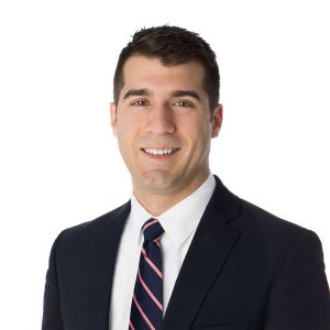 A smiling individual wearing a suit with a blue tie and a striped shirt against a white background.
