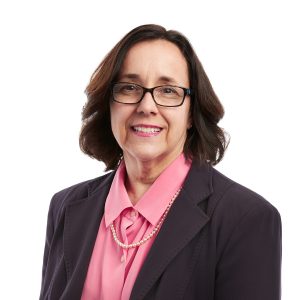 A professional woman wearing glasses, a pink blouse, and a dark blazer is smiling at the camera.