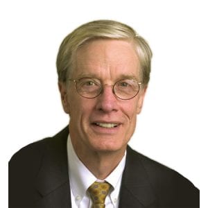 A smiling individual wearing glasses, a suit, and a tie with a plain background.