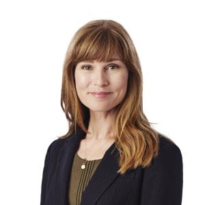 A woman with shoulder-length hair and bangs is smiling and wearing a dark blazer over a patterned blouse against a light background.
