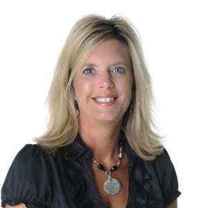 A smiling woman with shoulder-length blonde hair, wearing a black shirt and a silver necklace, is posing against a white background.