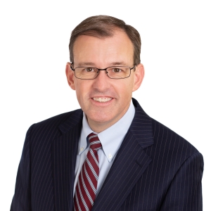A man in a business suit with a striped tie is smiling against a white background.