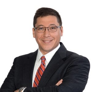 A smiling individual wearing glasses, a dark suit, and a red striped tie poses against a white background.
