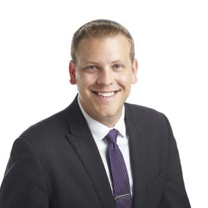 A smiling person wearing a dark suit and a purple tie against a white background.