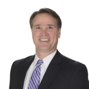 A smiling individual with a navy blue suit, white shirt, and purple-striped tie against a neutral background.