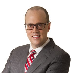 A man wearing glasses, a gray suit, and a red striped tie is smiling at the camera.