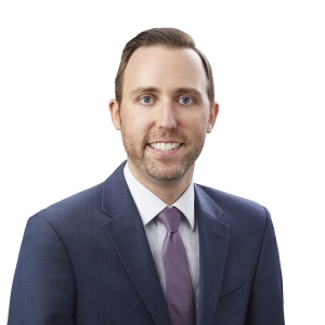 A smiling person wearing a professional suit and purple tie against a plain white background.