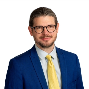A man with a light stubble is wearing a dark suit, a white shirt, and a yellow tie.