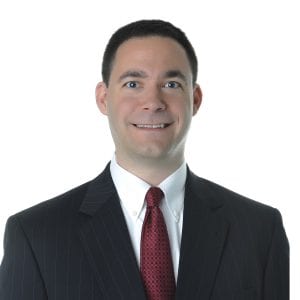 A smiling man wearing a suit with a pinstripe jacket, white shirt, and red tie.