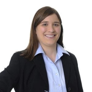 A person wearing a black blazer over a light blue collared shirt is smiling against a white background.