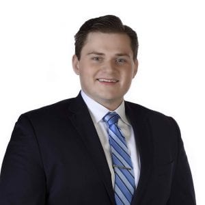 A person is wearing a dark suit, a light blue striped tie, and has a friendly smile.