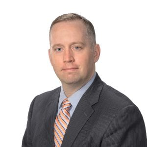A person wearing a suit with a striped tie poses for a professional portrait against a neutral background.