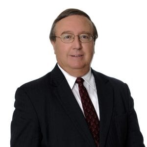 A man wearing a suit and tie with glasses is smiling against a plain background.