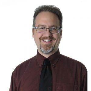 A smiling individual with glasses wearing a burgundy shirt and a black tie against a white background.